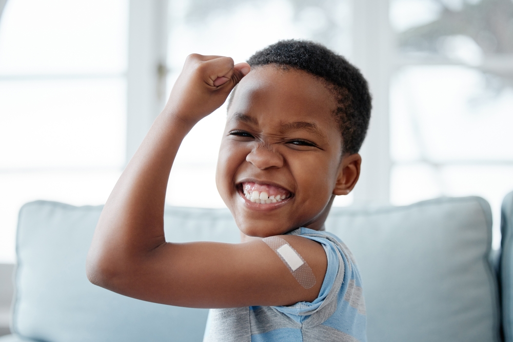 A smiling boy flexes his arm, showing off a bandage from a vaccination