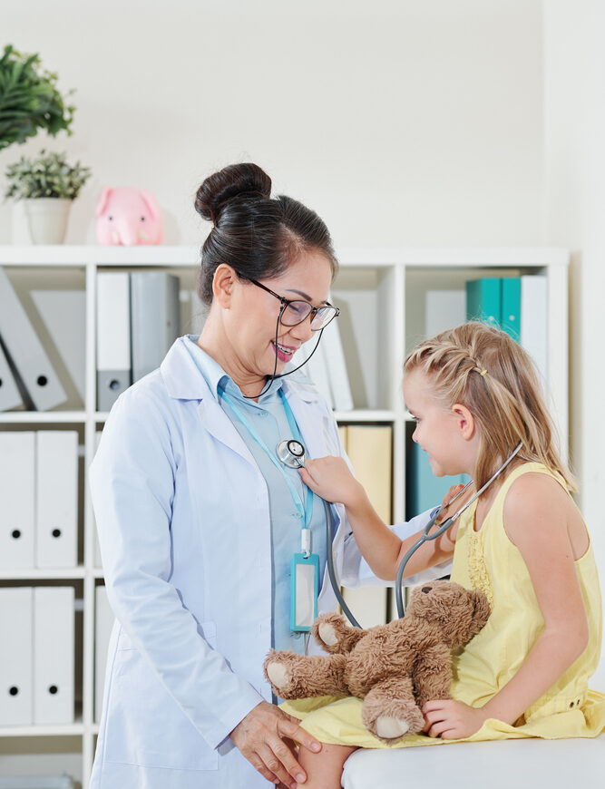 A pediatrician listens to a young girl's heartbeat with a stethoscope while she holds a teddy bear
