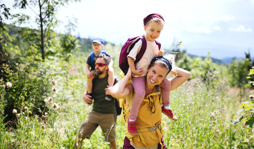 A mother and father carry their two young children on their shoulders while hiking a grassy trail