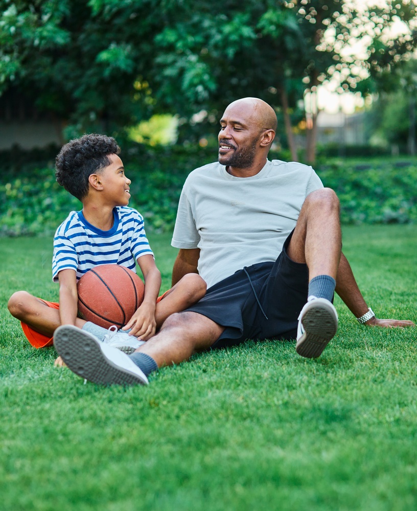 A father and young son sit together on the grass smiling, with a basketball between them