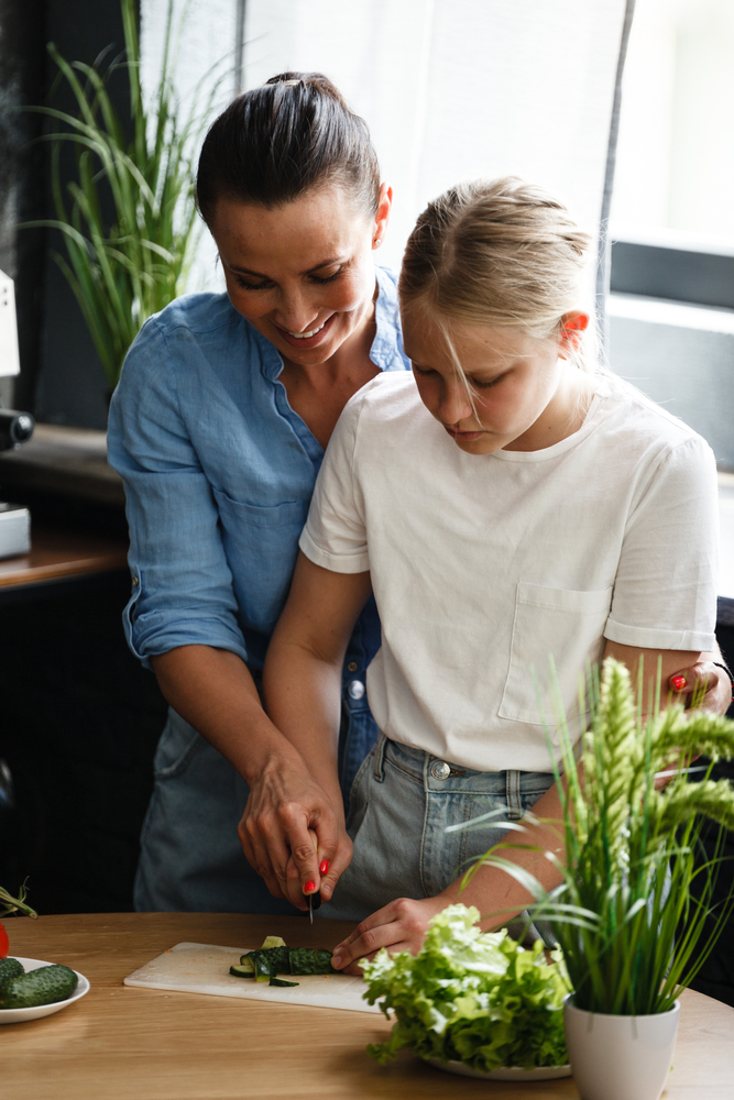 A mother helps her daughter cut cucumbers at a kitchen counter