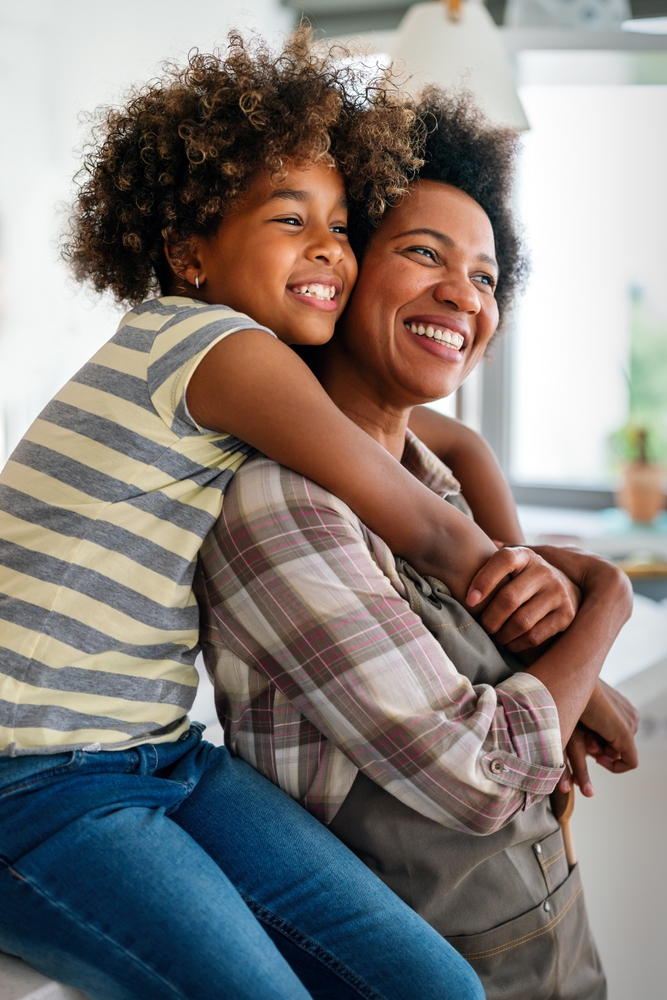 A mother and daughter with curly hair smile and embrace in a bright kitchen