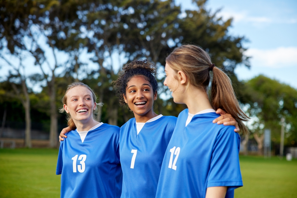 Three smiling teen girls in blue sports jerseys standing arm in arm outdoors
