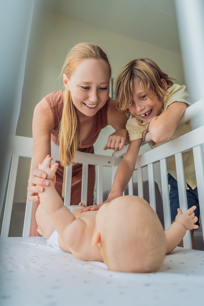 A mother and older brother smiling over a baby lying in a crib