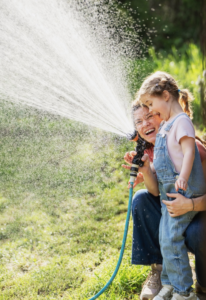 A mother and daughter laughing together while playing with a garden hose